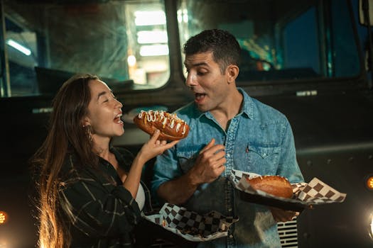 A couple happily enjoying hot dogs outside a food truck at night.