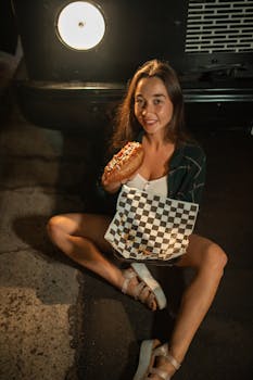 Young woman sitting near a food truck holding a hot dog, smiling at night.