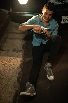 晚上，一名男子坐在街上，快乐地对着汽车前灯吃三明治