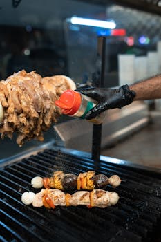 Gloved chef's hand seasoning kebabs on a grill indoors with a shaker.
