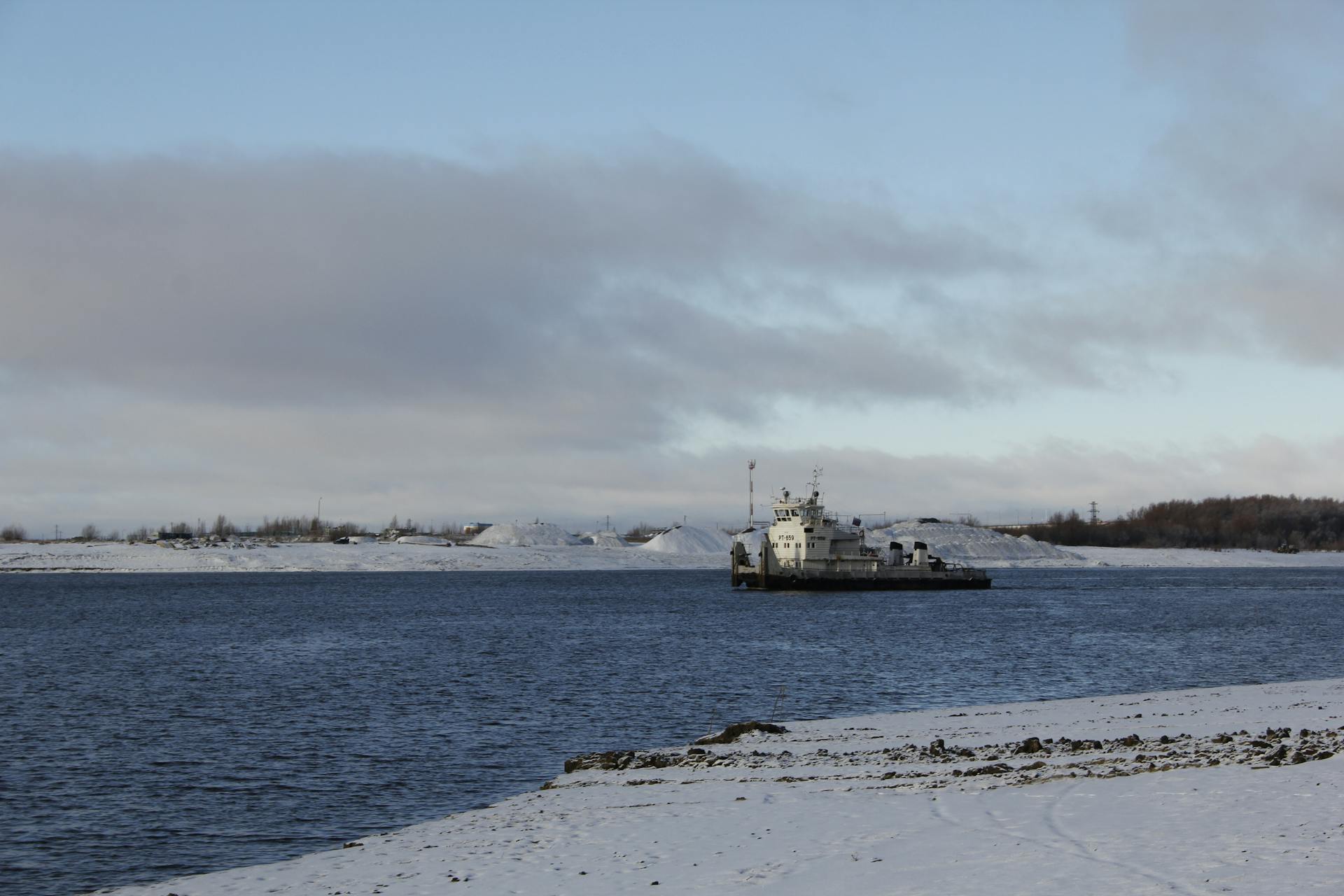 A peaceful winter scene with a ship on a snowy river under a cloudy sky.