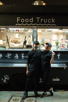 A man and woman stand in front of a food truck, offering street food at night.