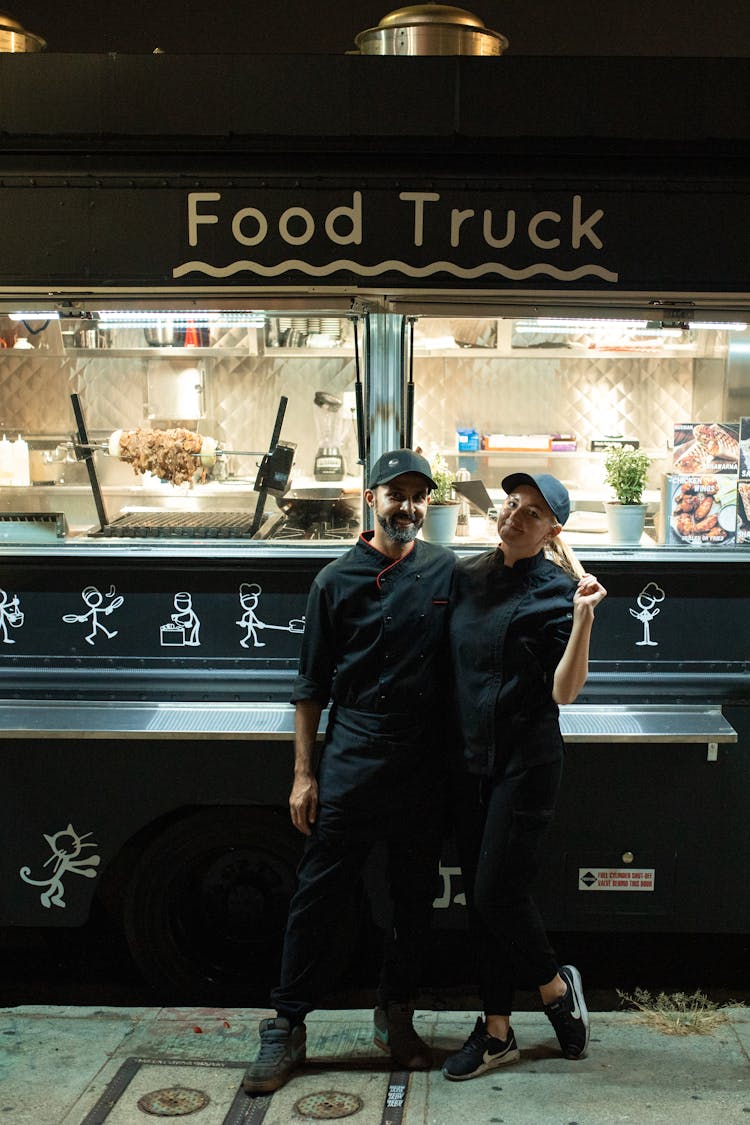 Man And Woman In Blue Button Up Shirt And Black Pants Standing Beside Food Truck