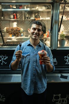 A man smiling while holding street food and a fork at an outdoor food truck during nighttime.