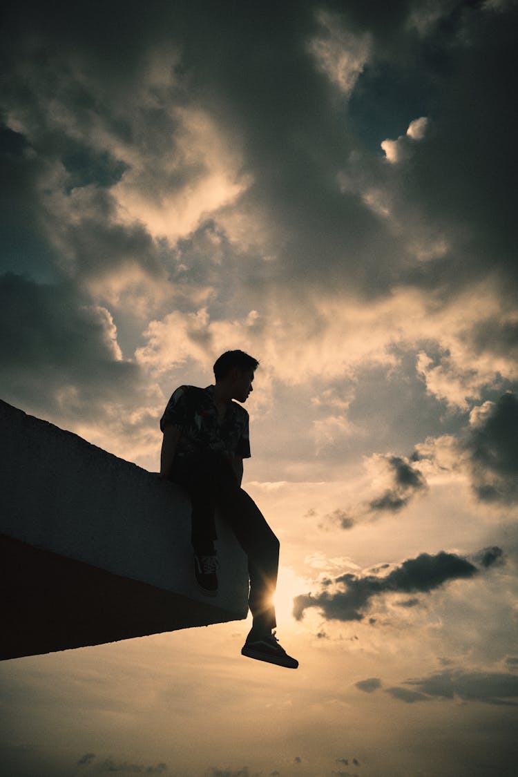 Unrecognizable Man Sitting On Rooftop Edge Against Cloudy Sundown Sky