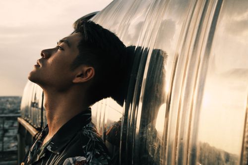Free Side view of sad young Asian guy with dark hair leaning on metal mirrored wall while relaxing on building rooftop at sundown Stock Photo