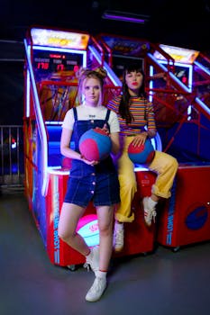 Two young women playing basketball arcade games with neon lights.