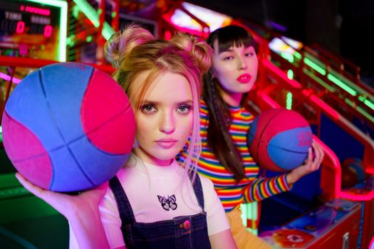 Two fashionable women holding basketballs in a vibrant, neon-lit amusement arcade.