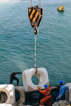 A team of workers uses a crane to move large concrete blocks near the sea shore.