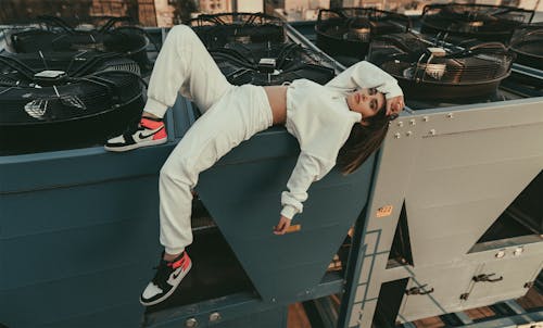 Full body of stylish female with long hair looking at camera while lying with bent leg on edge of compressor air conditioner on roof