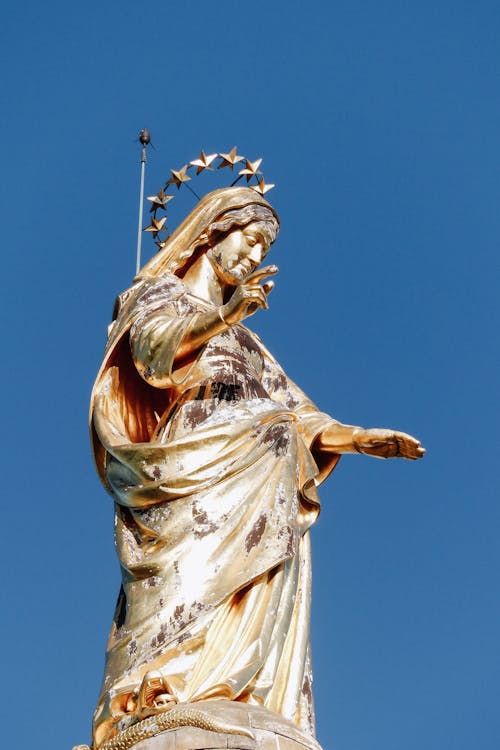 Low angle of famous golden sculpture of Virgin Mary in crown of stars placed on pillar against blue sky outside