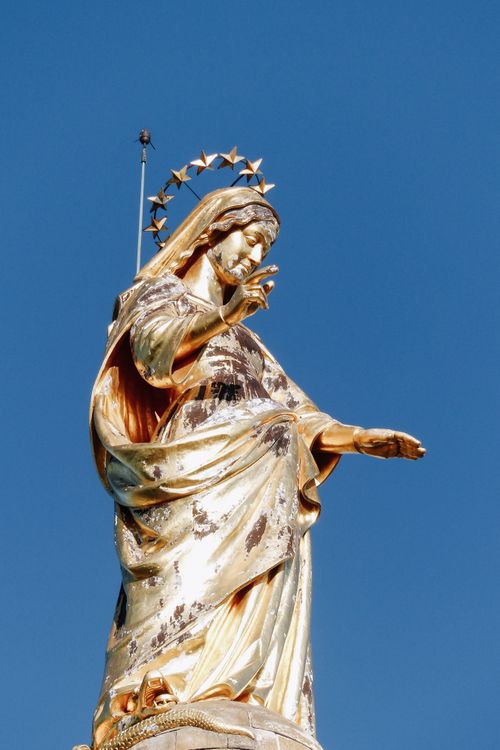 Free Low angle of famous golden sculpture of Virgin Mary in crown of stars placed on pillar against blue sky outside Stock Photo