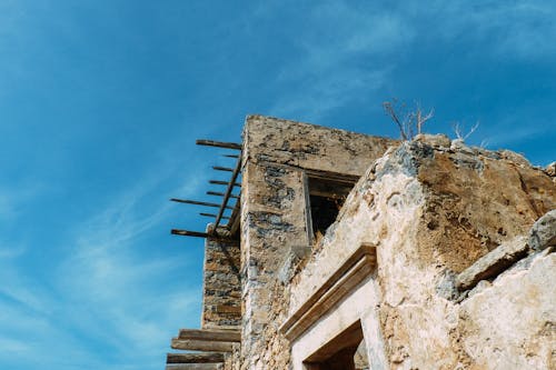 From below of damaged stone building with window holes and timber on roof against blue sky in countryside in sunlight