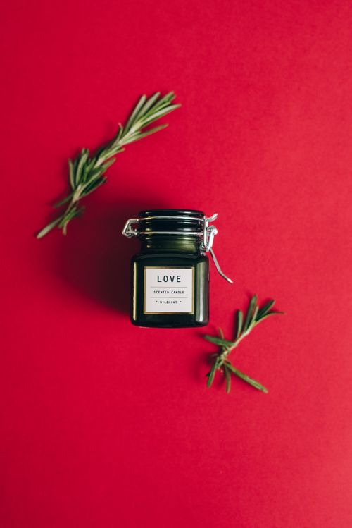 Free Glass jar scented candle labeled 'Love' adorned with rosemary sprigs on a red background. Stock Photo