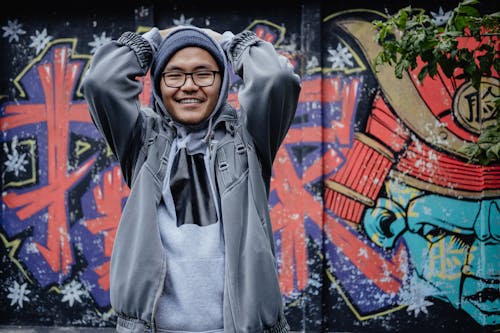 Free Young happy ethnic guy in trendy clothes with hands behind head looking at camera near colorful graffiti wall Stock Photo