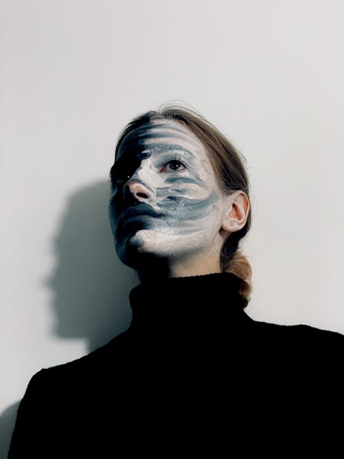From below of calm strange female with paints on face wearing black turtleneck looking away while standing against white background