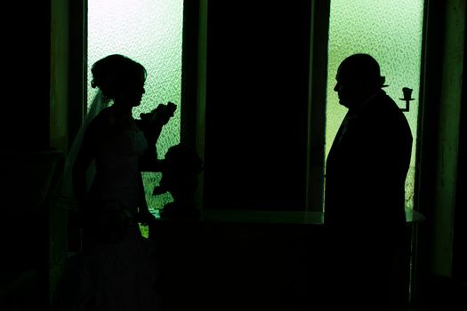 Artistic silhouette of a bride and groom facing each other in a dark room with green windows.