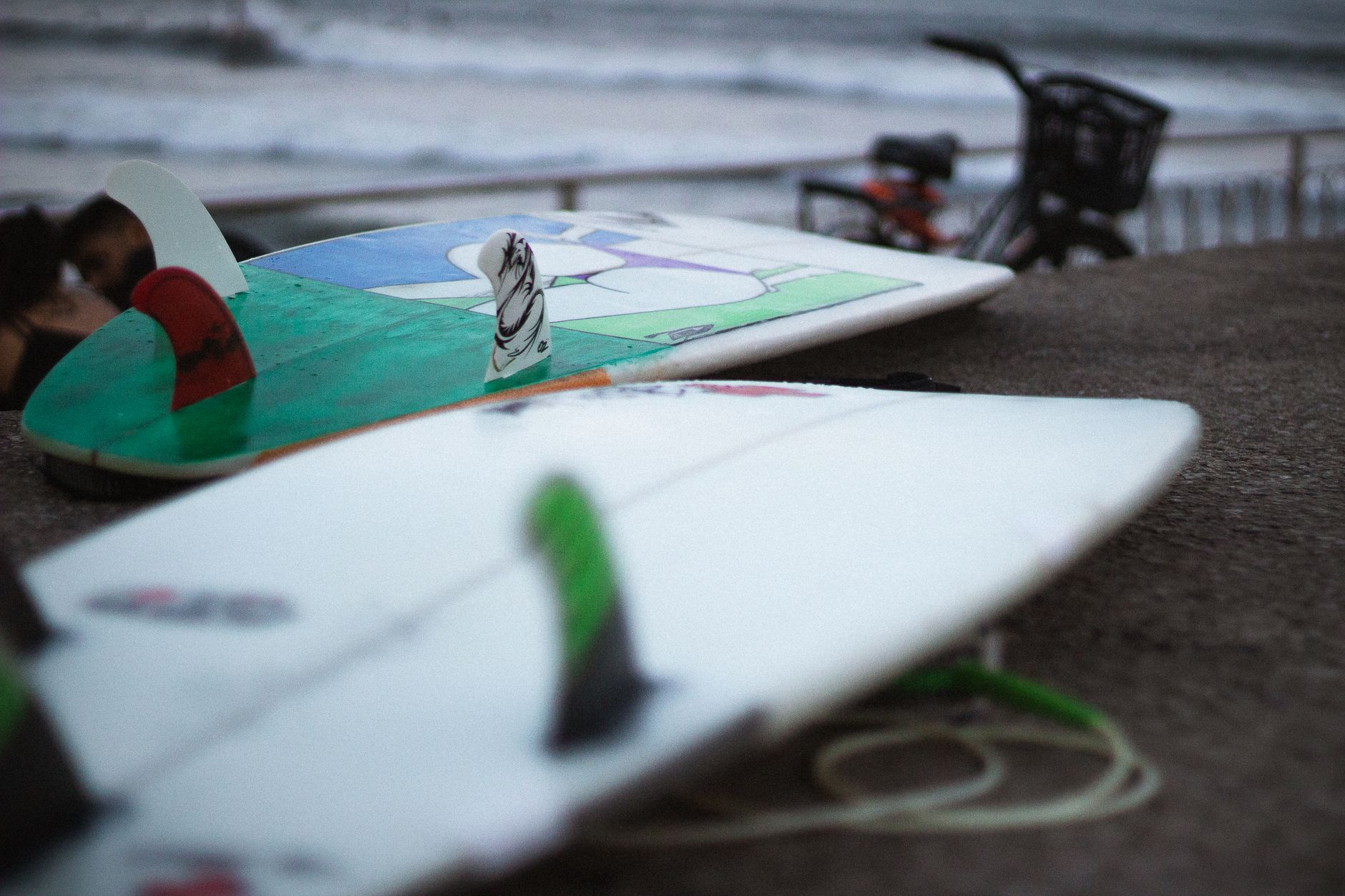 Close-up of surfboards on a beachside with waves and bicycle in the background.