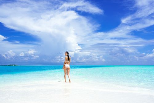 Free A woman in a bikini stands on a pristine beach under a vibrant blue sky, with turquoise water and white sand. Stock Photo