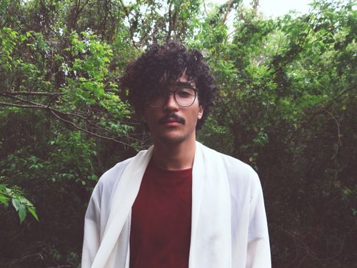 Free Portrait of a young man with curly hair and glasses wearing a red shirt in a forested setting. Stock Photo