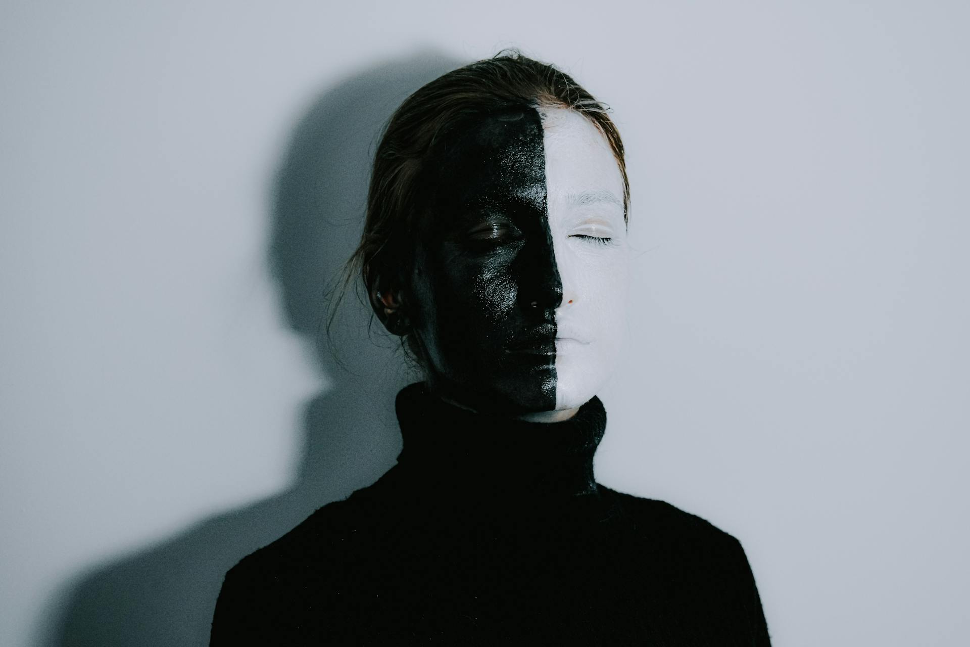 Mysterious female with face painted half in white and half in black colors standing with closed eyes against light background