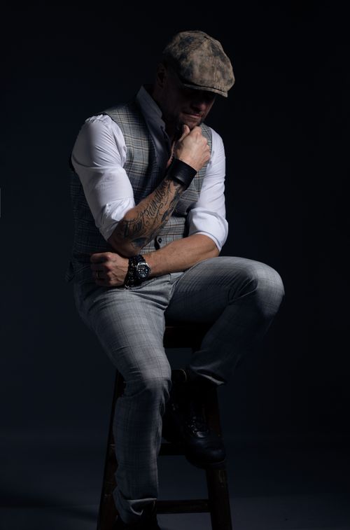 Free A man with arm tattoo posing thoughtfully in a studio shoot, wearing a flat cap and stylish outfit. Stock Photo