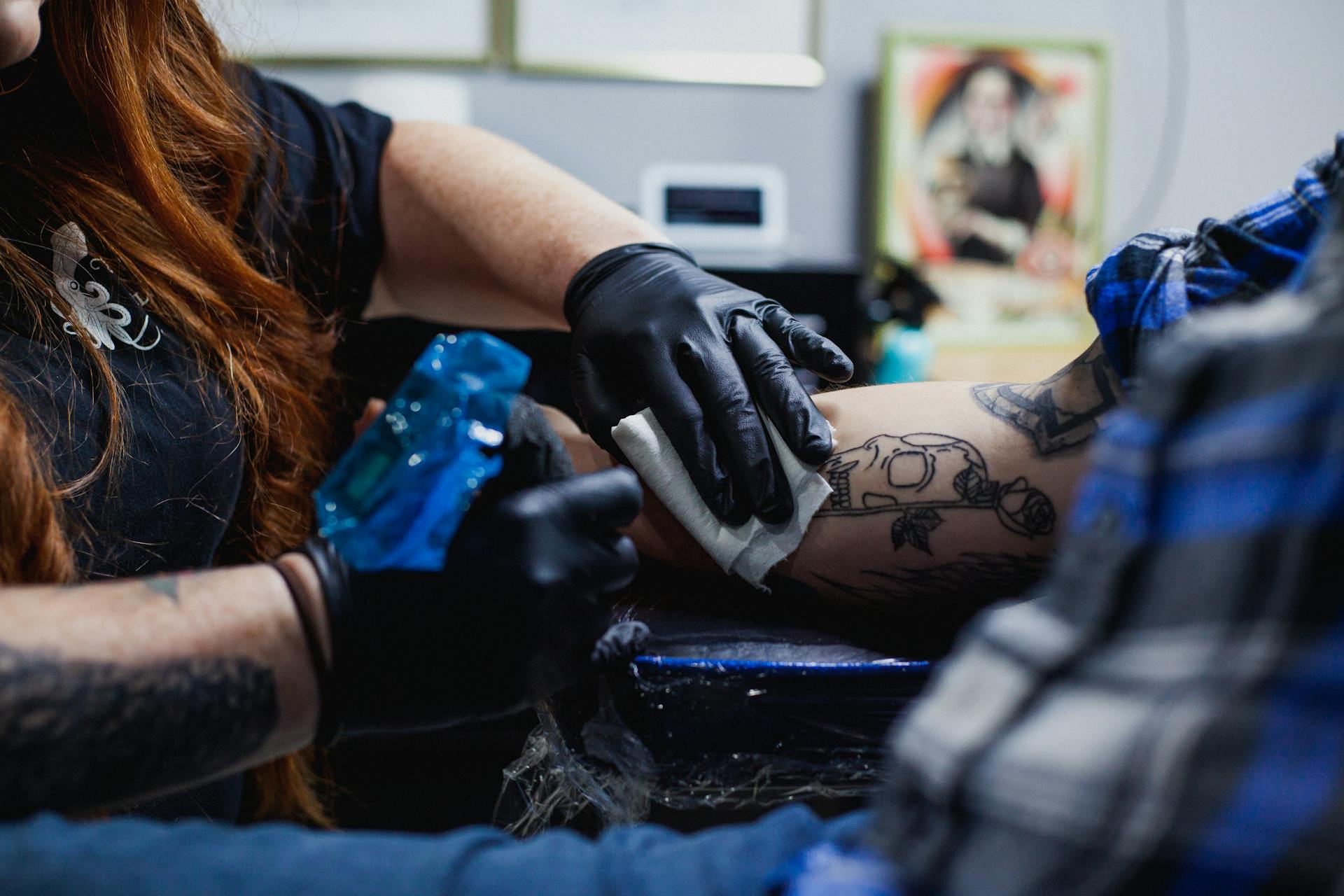 Faceless woman in black gloves doing tattoo with gun on arm of man in salon