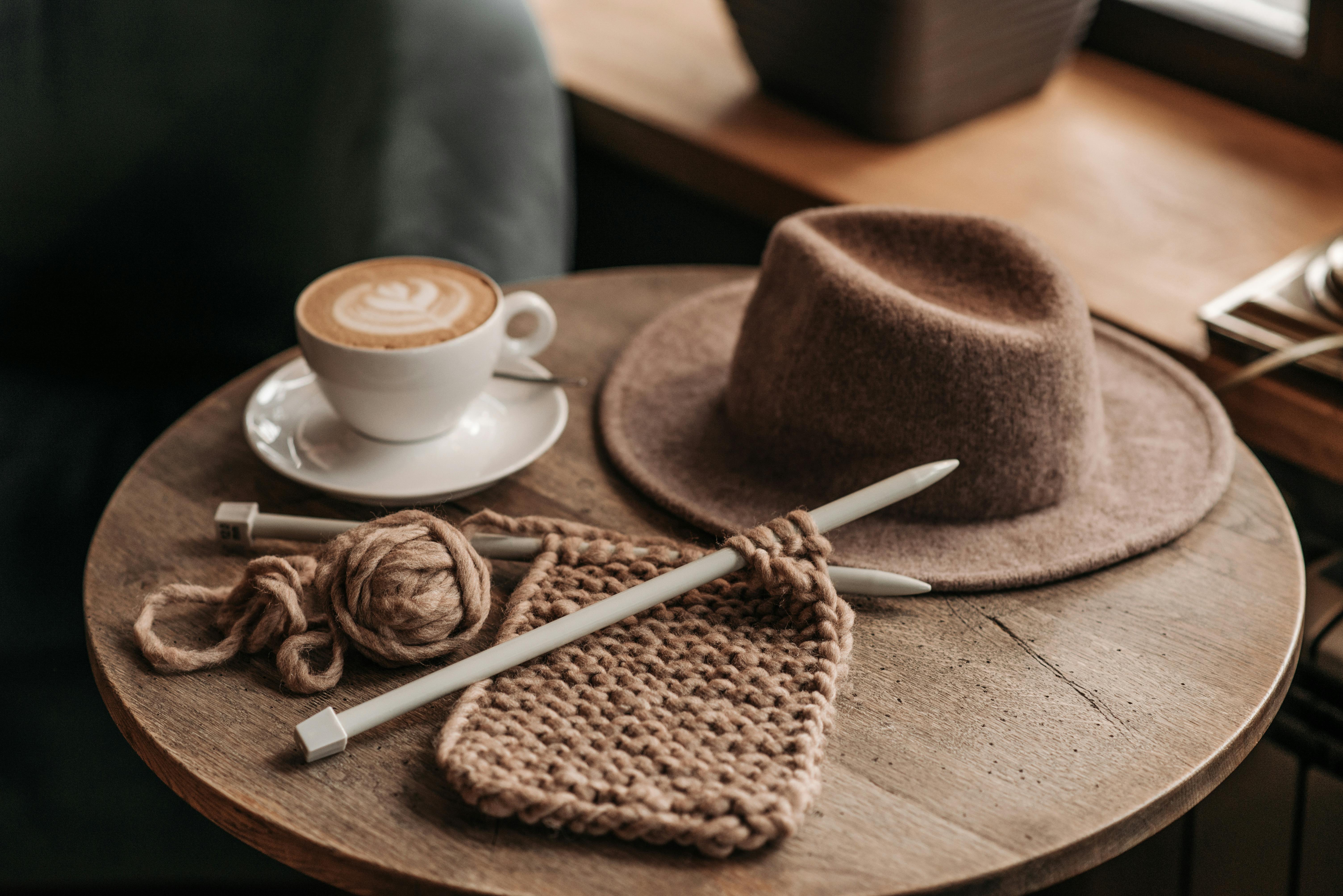 Close-up of a cup of coffee, knitting, and hat on a rustic table indoors.