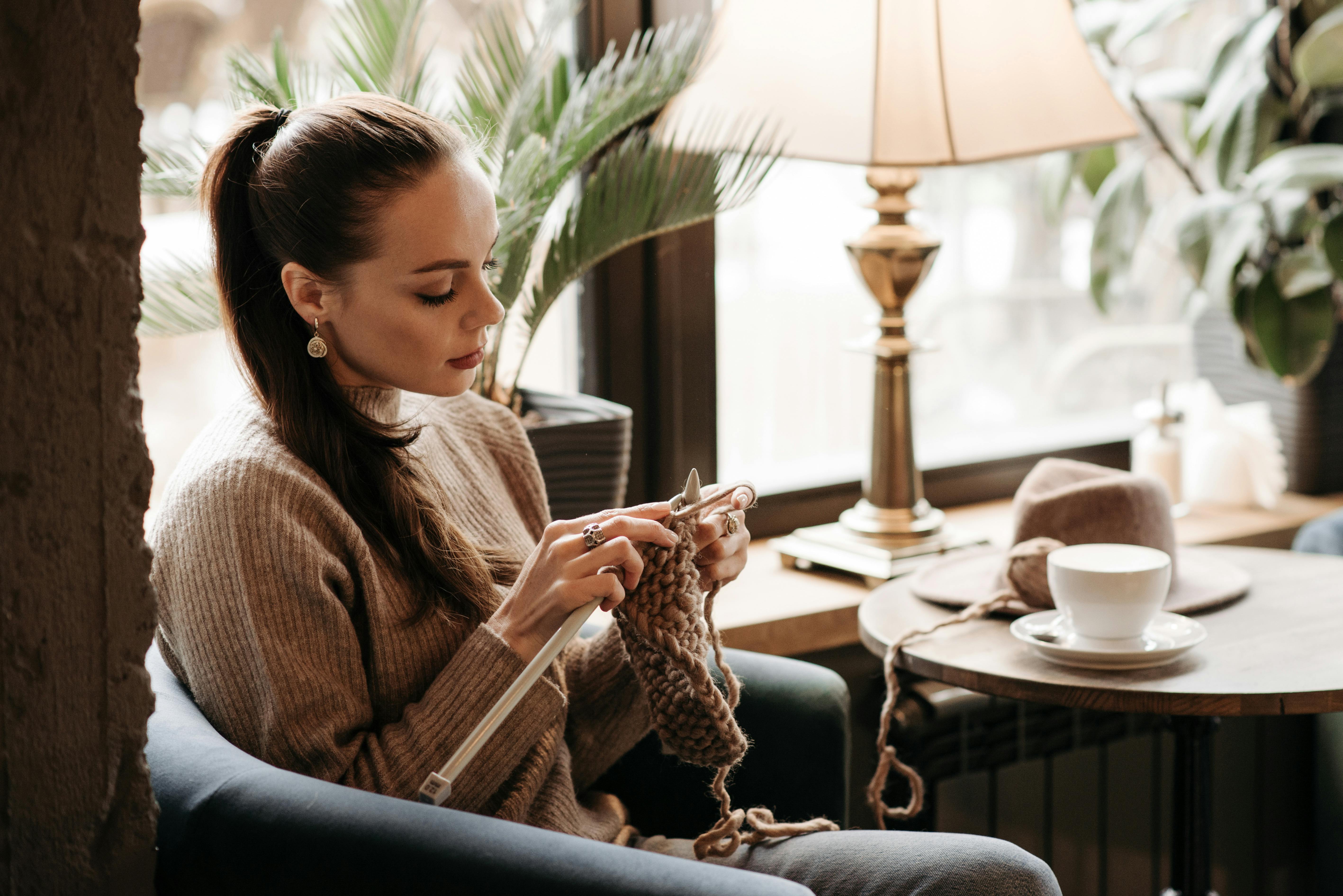 Woman knitting near a window with yarn and a cup of coffee on a round table, enjoying a relaxing indoor setting.