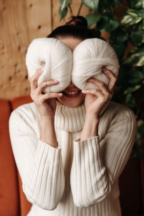 Free Smiling woman holds white yarn rolls over her eyes indoors. Cozy and playful vibe. Stock Photo