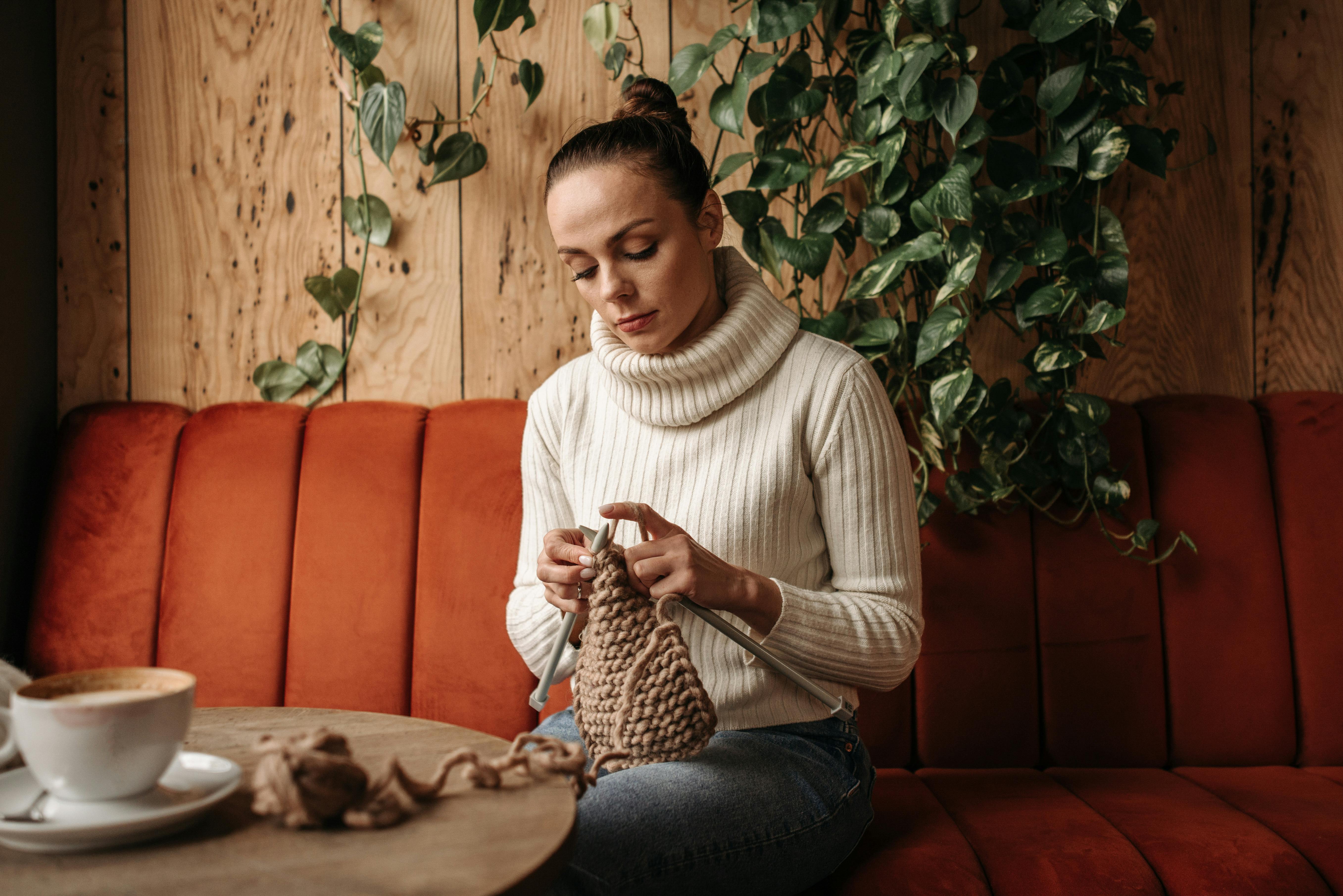 A woman in a turtleneck sweater knits in a cozy cafe setting with plants and a warm ambiance.