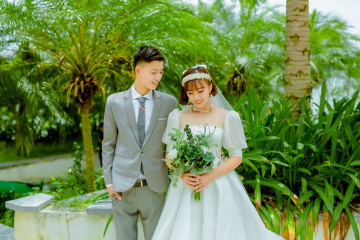 A joyful Asian couple in bridal attire posing outdoors amidst lush greenery.