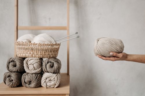 Free Hand holding a ball of yarn next to a basket with knitting needles and yarn rolls on a wooden table. Stock Photo