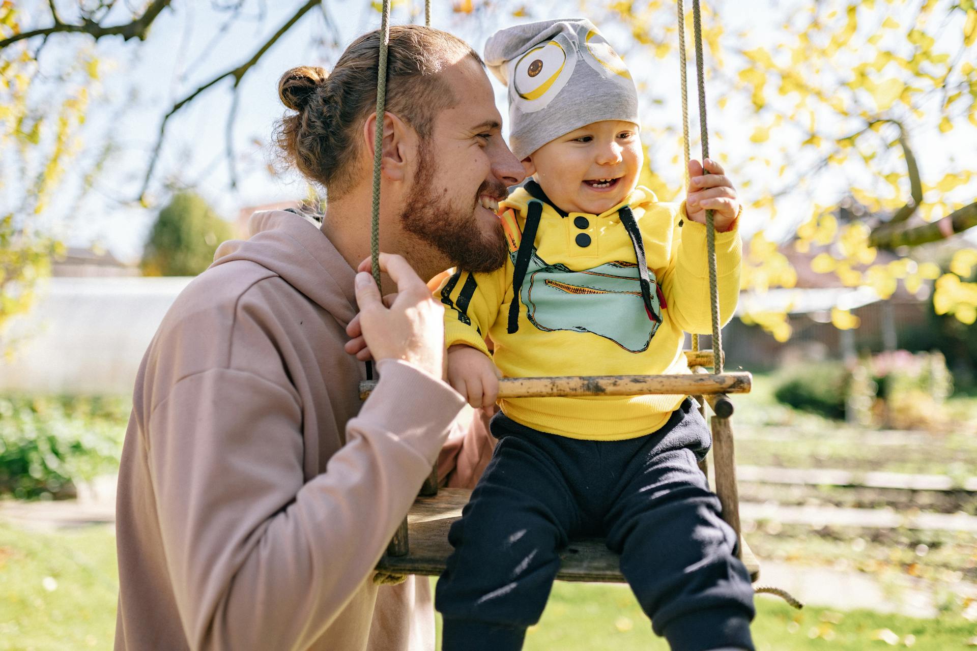A father joyfully interacts with his toddler on a swing during a sunny fall day outdoors.
