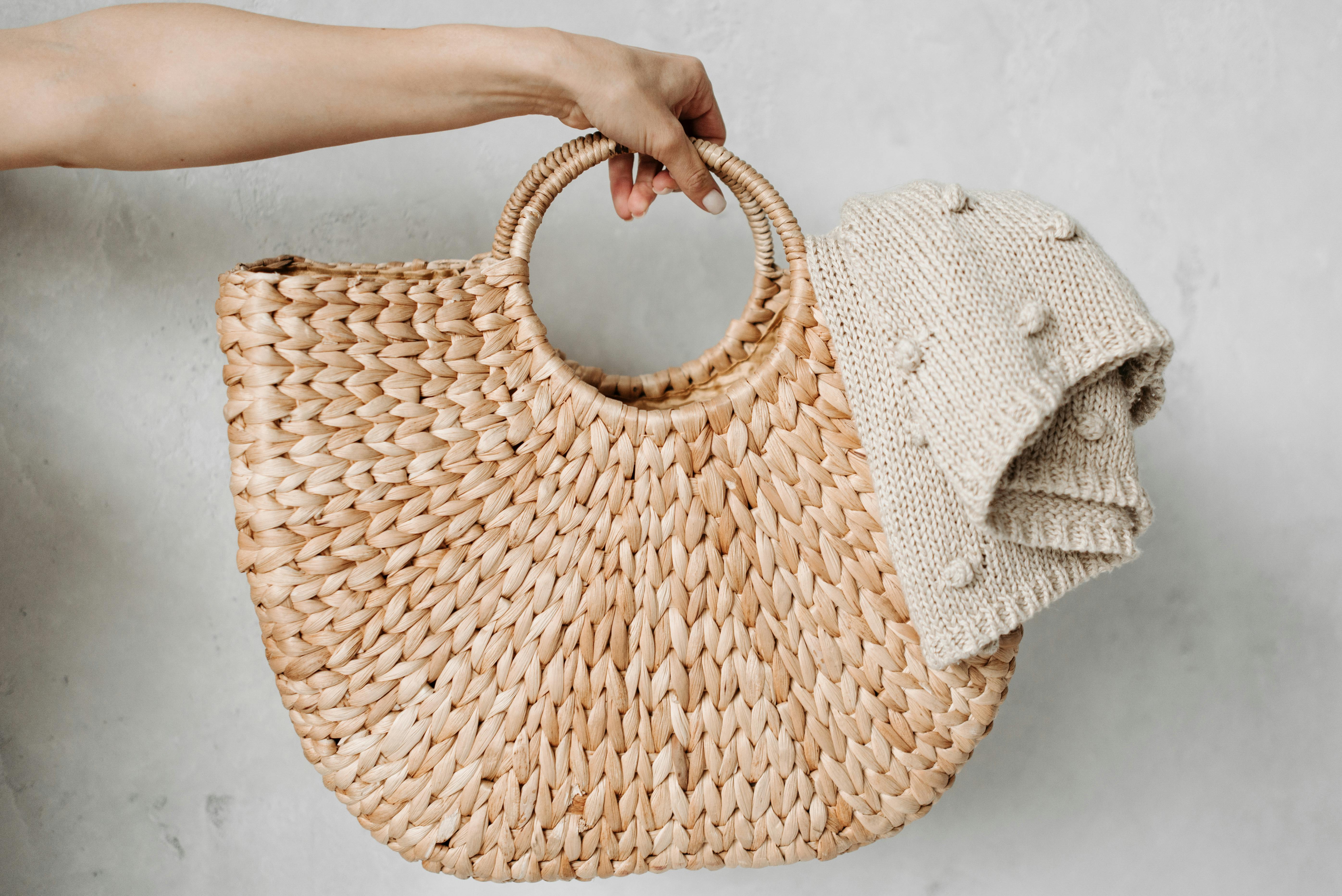 A hand holding a stylish woven bag with a knitted sweater draped over it.