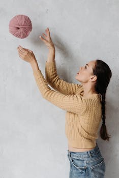 A woman in a cozy sweater tosses a large pink yarn ball indoors against a textured wall.