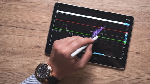 Close-up of a hand using a stylus on a digital trading app on a tablet indoors.