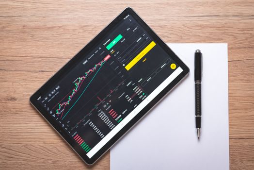 Overhead view of a digital tablet displaying stock market graphs and data on a wooden desk.