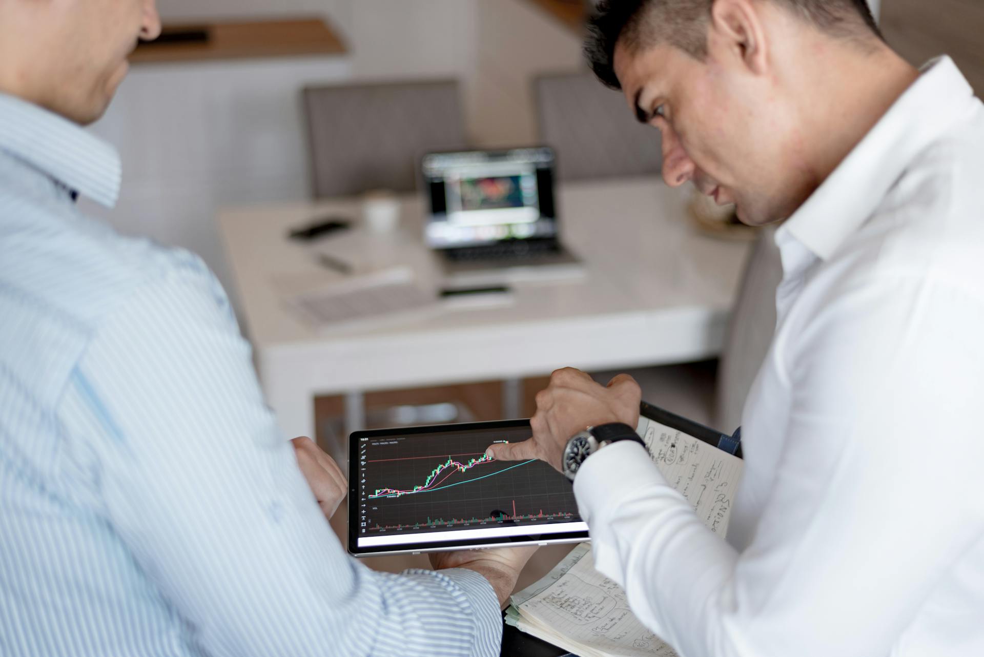 Two businessmen discuss stock market trends using a tablet with visible graphs.