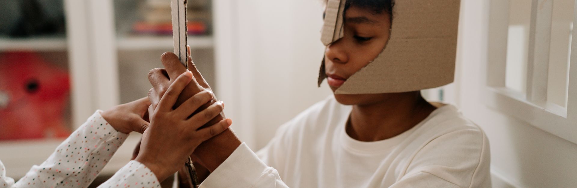 A young child dressed in a cardboard helmet engages in imaginative play with a handmade sword indoor