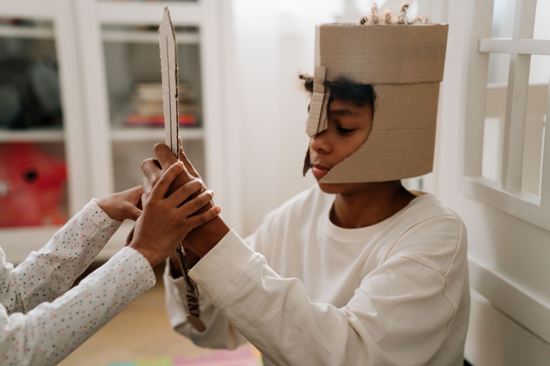 A young child dressed in a cardboard helmet engages in imaginative play with a handmade sword indoor