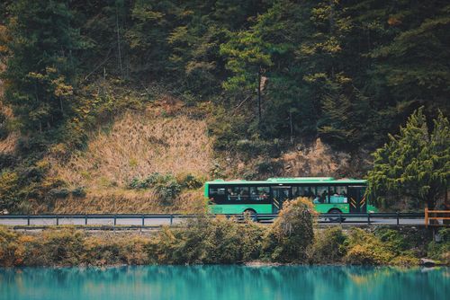 Free A green bus travels along a forest road beside a calm, turquoise water body, offering a serene travel scene. Stock Photo