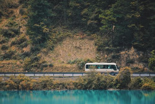 Free A white bus travels by a scenic forested road along a turquoise river, capturing a tranquil journey. Stock Photo