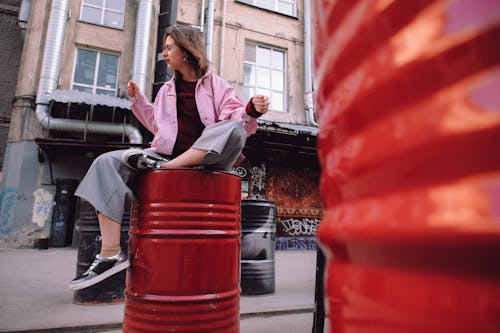 Low angle full body of peaceful female in stylish casual clothes sitting on metal barrel