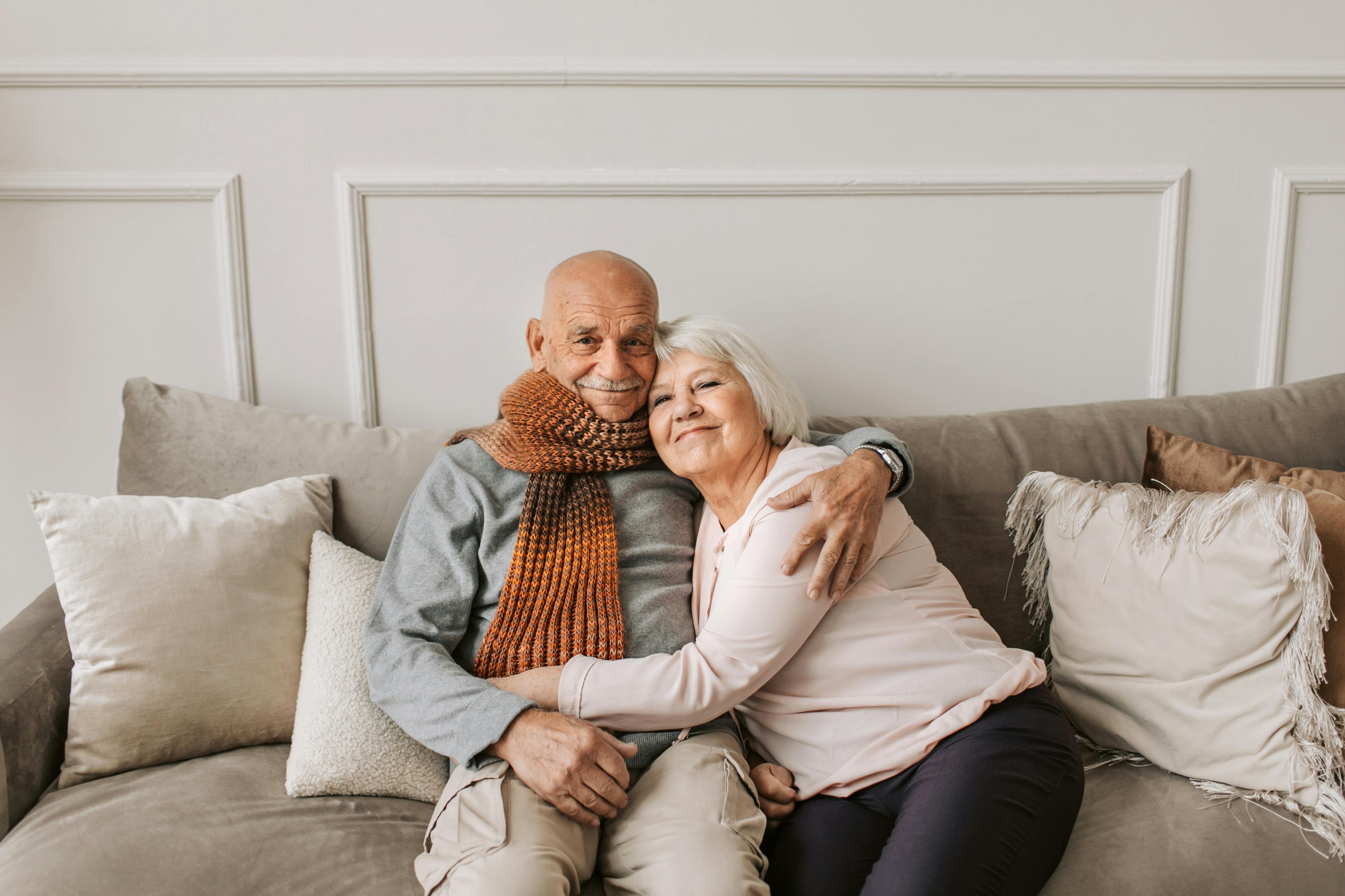 Elderly couple embracing on a sofa, smiling warmly. Neutral setting with pillows.