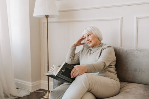 Free Senior woman sitting on a couch reading a book, with a thoughtful expression. Stock Photo