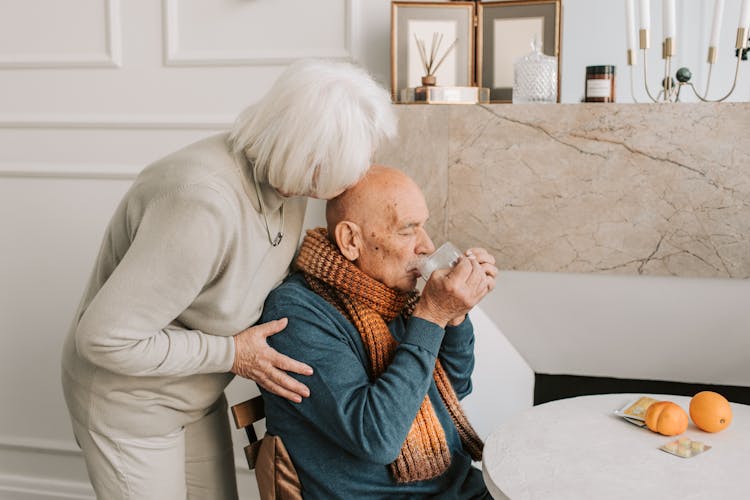 Elderly Man In Blue Sweater Drinking Medicine Beside An Elderly Woman In Gray Sweater