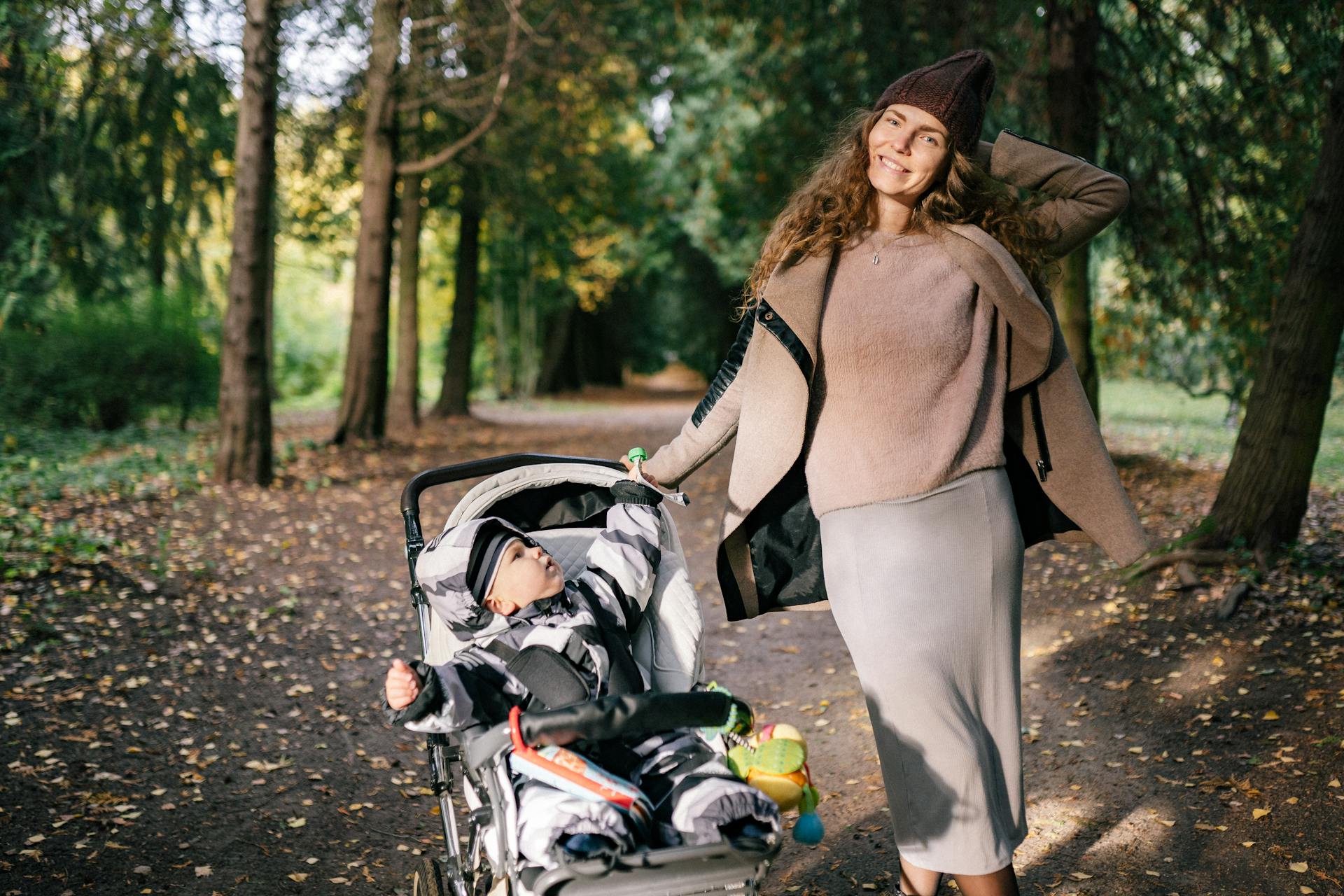 A joyful mother poses with her baby in a stroller while enjoying a stroll in a beautiful autumn park.