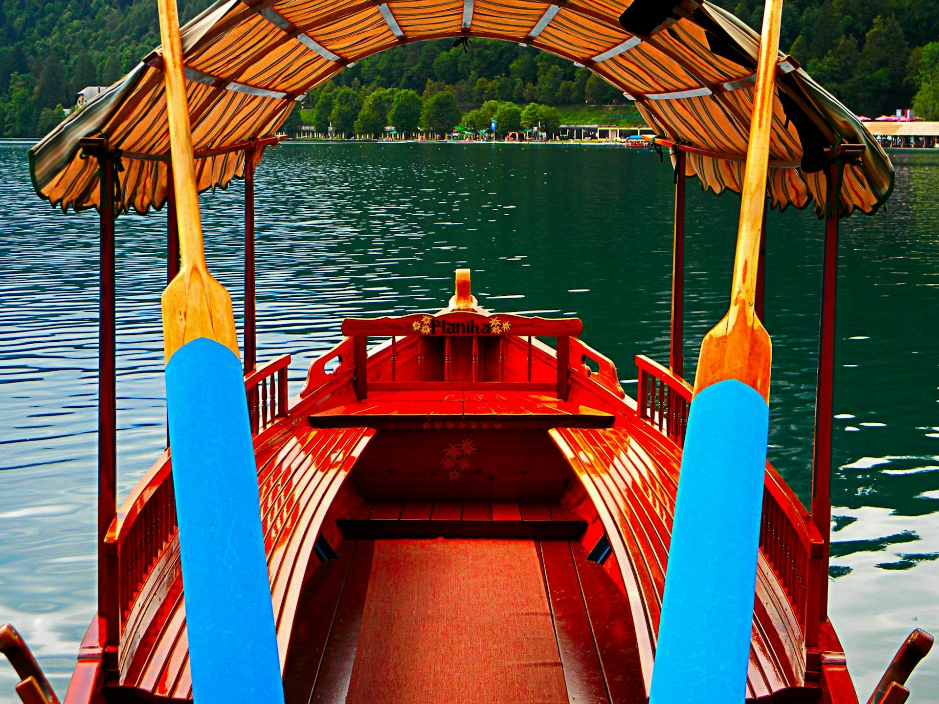 Picture of a traditional wooden boat with blue oars on Lake Bled, Slovenia, showcasing serene waters and lush surroundings.