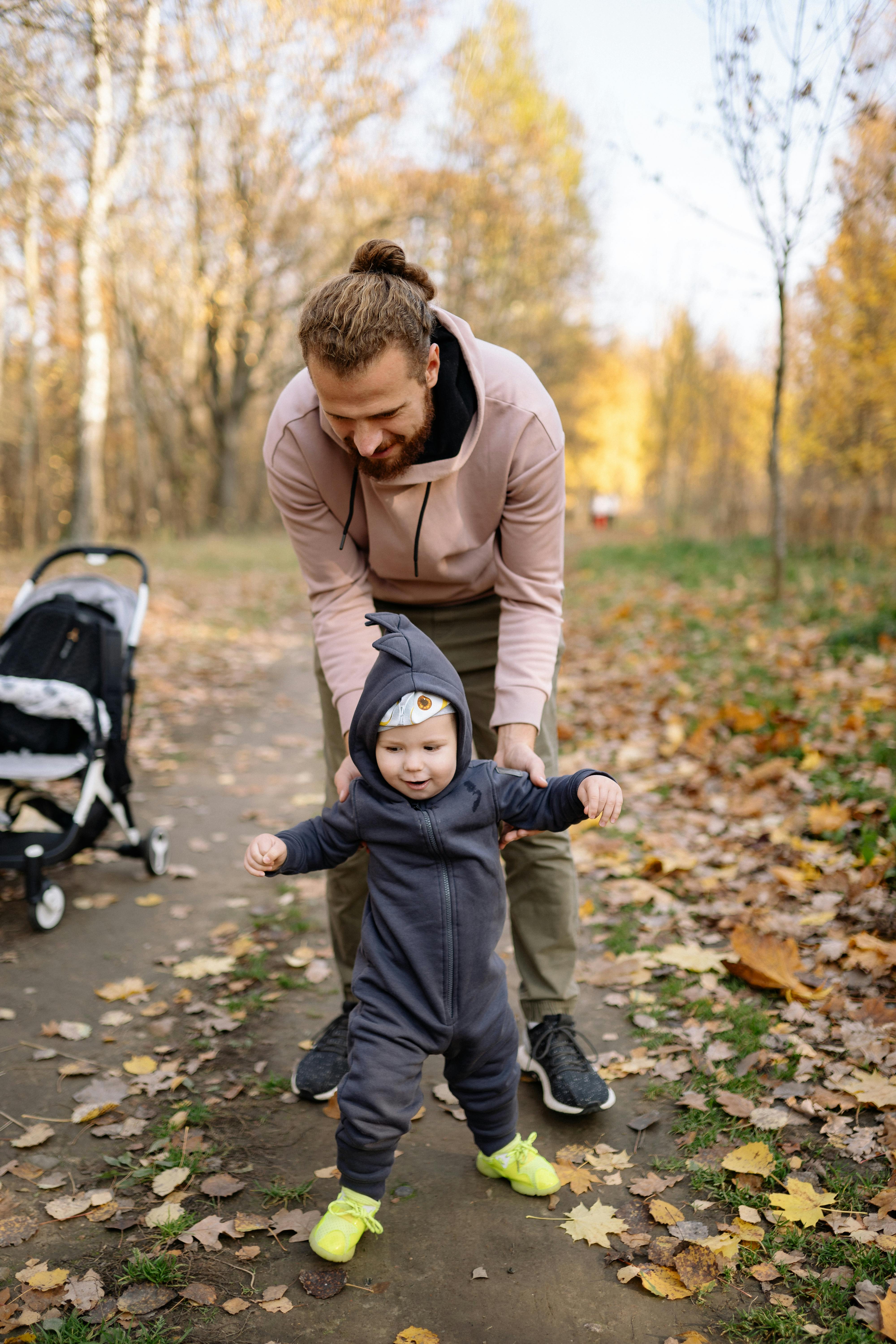 A father helps his child learn to walk on a leafy path during autumn in the park.